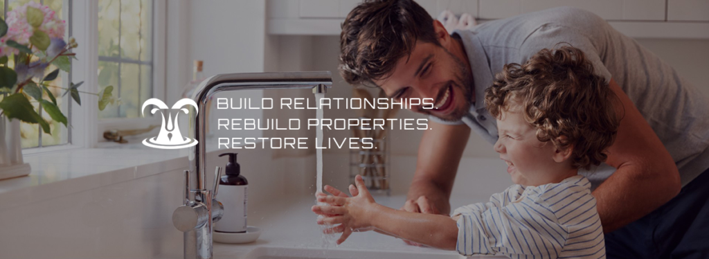 Man leaning on counter smiling at toddler son while son washes his hands at the kitchen sink. Text overlaying image says "Build Relationships. Rebuild Properties. Restore Lives."