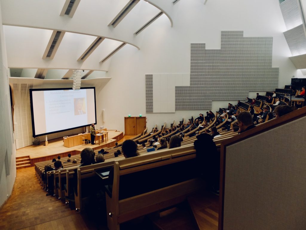 Stadium-style lecture hall with students seated and professor teaching onstage with large projector screen.